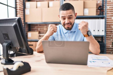 Foto de Hispanic man working at small business ecommerce with laptop screaming proud, celebrating victory and success very excited with raised arm - Imagen libre de derechos