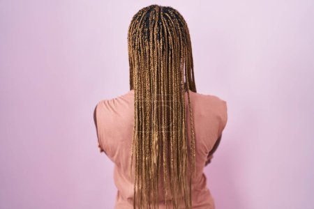Téléchargez les photos : Femme afro-américaine avec les cheveux tressés debout sur fond rose debout vers l'arrière regardant loin avec les bras croisés - en image libre de droit