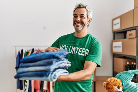 Téléchargez les photos : Moyen-âge homme aux cheveux gris volontaire souriant confiant tenant jeans pliés au centre de charité - en image libre de droit