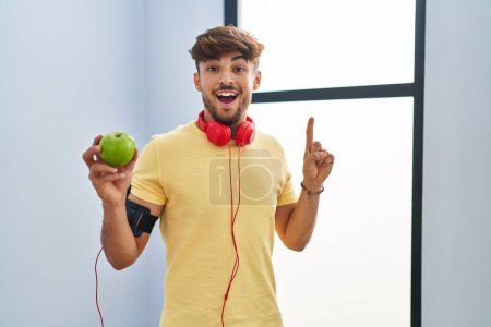 Photo for Arab man with beard wearing sportswear eating green apple smiling with an idea or question pointing finger with happy face, number one - Royalty Free Image