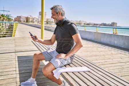 Téléchargez les photos : Homme hispanique du Moyen Âge portant des vêtements de sport en utilisant un smartphone au bord de la mer - en image libre de droit