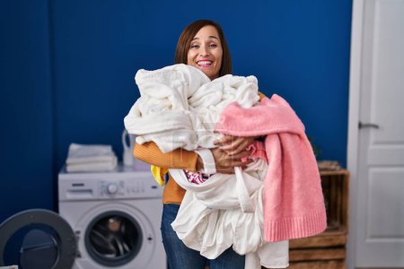 Foto de Middle age woman holding dirty laundry ready to put it in the washing machine smiling and laughing hard out loud because funny crazy joke. - Imagen libre de derechos