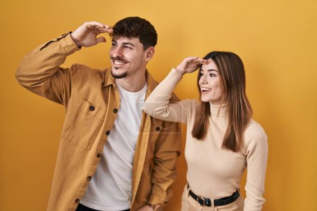 Photo for Young hispanic couple standing over yellow background very happy and smiling looking far away with hand over head. searching concept. - Royalty Free Image