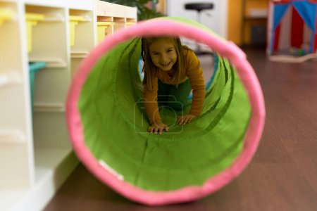 Foto de Adorable hispanic girl crawling inside tunnel toy at kindergarten - Imagen libre de derechos