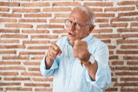 Photo for Senior man with grey hair standing over bricks wall ready to fight with fist defense gesture, angry and upset face, afraid of problem - Royalty Free Image
