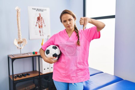 Photo for Young hispanic woman working at physiotherapy clinic holding football ball with angry face, negative sign showing dislike with thumbs down, rejection concept - Royalty Free Image