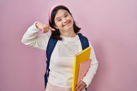 Téléchargez les photos : Woman with down syndrome wearing student backpack and holding books smiling cheerful showing and pointing with fingers teeth and mouth. dental health concept. - en image libre de droit
