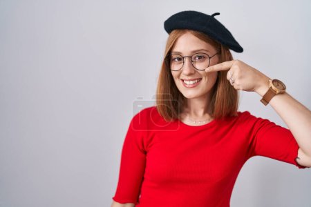 Téléchargez les photos : Young redhead woman standing wearing glasses and beret pointing with hand finger to face and nose, smiling cheerful. beauty concept - en image libre de droit