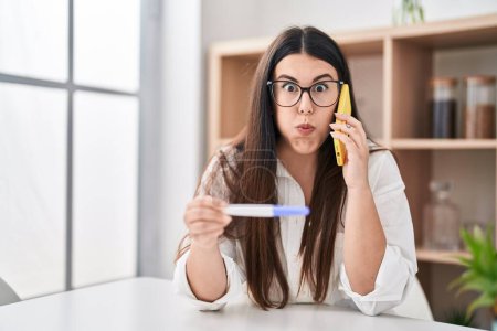 Photo for Young brunette woman holding pregnancy test result speaking on the phone puffing cheeks with funny face. mouth inflated with air, catching air. - Royalty Free Image