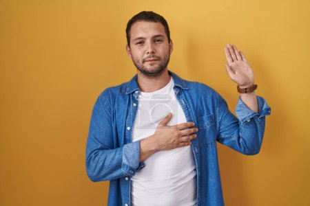 Photo for Hispanic man standing over yellow background swearing with hand on chest and open palm, making a loyalty promise oath - Royalty Free Image