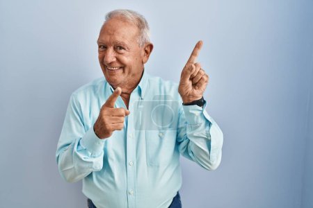 Foto de Senior man with grey hair standing over blue background smiling and looking at the camera pointing with two hands and fingers to the side. - Imagen libre de derechos