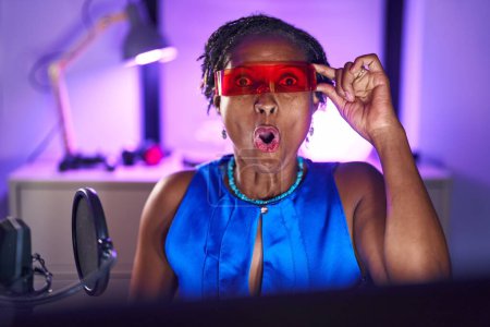 Téléchargez les photos : African woman with dreadlocks playing video games wearing virtual reality glasses scared and amazed with open mouth for surprise, disbelief face - en image libre de droit