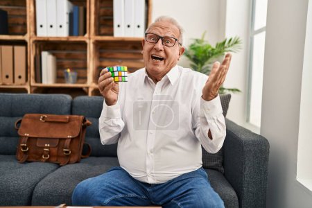 Téléchargez les photos : Senior therapist with grey hair playing colorful puzzle cube intelligence game celebrating achievement with happy smile and winner expression with raised hand - en image libre de droit