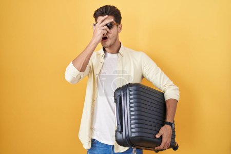 Téléchargez les photos : Young hispanic man holding suitcase going on summer vacation peeking in shock covering face and eyes with hand, looking through fingers with embarrassed expression. - en image libre de droit
