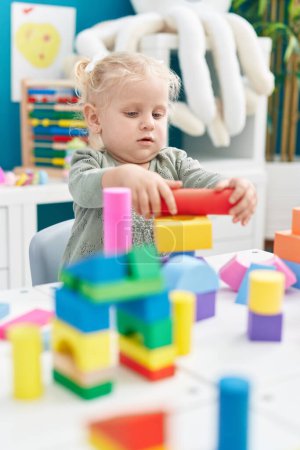 Foto de Adorable chica rubia jugando con bloques de construcción sentado en la mesa en el jardín de infantes - Imagen libre de derechos
