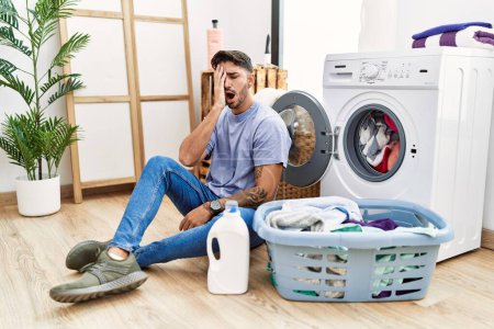 Téléchargez les photos : Young hispanic man putting dirty laundry into washing machine yawning tired covering half face, eye and mouth with hand. face hurts in pain. - en image libre de droit