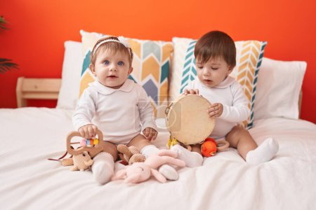 Foto de Dos adorables bebés sosteniendo pandereta sentados en la cama en el dormitorio - Imagen libre de derechos