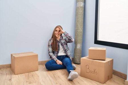 Téléchargez les photos : Young blonde woman sitting on the floor moving to a new home doing ok gesture with hand smiling, eye looking through fingers with happy face. - en image libre de droit
