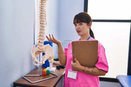 Photo for Young beautiful hispanic woman physiotherapist standing by anatomical model of spinal column speaking at rehab clinic - Royalty Free Image