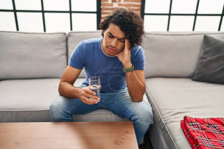 Téléchargez les photos : Jeune homme hispanique mal à l'aise boire de l'eau assis sur le canapé à la maison - en image libre de droit