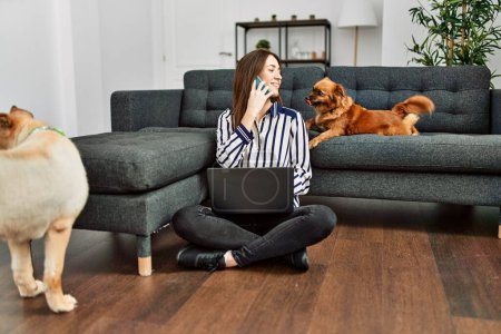 Téléchargez les photos : Jeune femme hispanique utilisant un ordinateur portable et parlant sur le smartphone assis sur le sol avec des chiens à la maison - en image libre de droit
