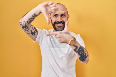 Foto de Young hispanic man with tattoos standing over yellow background smiling making frame with hands and fingers with happy face. creativity and photography concept. - Imagen libre de derechos
