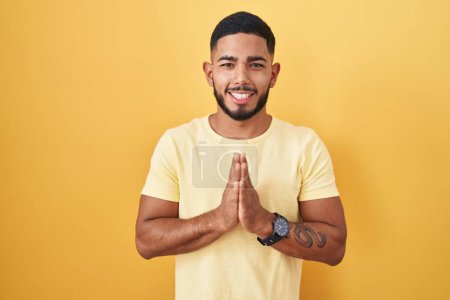 Photo for Young hispanic man standing over yellow background praying with hands together asking for forgiveness smiling confident. - Royalty Free Image