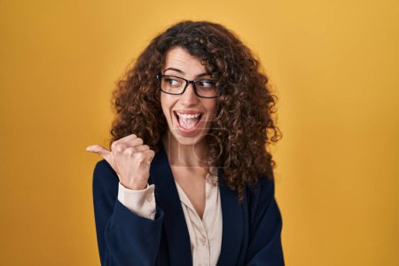 Foto de Hispanic woman with curly hair standing over yellow background smiling with happy face looking and pointing to the side with thumb up. - Imagen libre de derechos