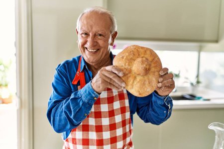 Foto de Hombre mayor sonriendo confiado sosteniendo pan casero en la cocina - Imagen libre de derechos