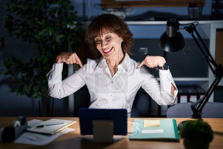 Téléchargez les photos : Middle age woman working at the office at night looking confident with smile on face, pointing oneself with fingers proud and happy. - en image libre de droit