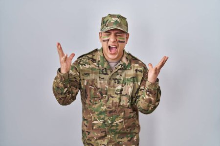 Foto de Hispanic young man wearing camouflage army uniform celebrating mad and crazy for success with arms raised and closed eyes screaming excited. winner concept - Imagen libre de derechos