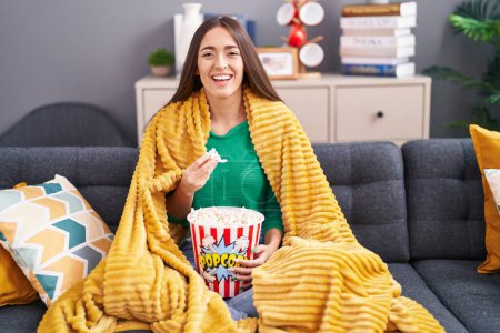 Foto de Young hispanic woman eating snack sitting on the sofa at home smiling and laughing hard out loud because funny crazy joke. - Imagen libre de derechos