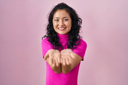Foto de Joven mujer asiática de pie sobre fondo rosa sonriendo con las palmas de las manos juntas recibiendo o dando gesto. retención y protección - Imagen libre de derechos