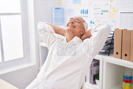Téléchargez les photos : Moyen âge homme aux cheveux gris travailleur d'affaires détendu avec les mains sur la tête au bureau - en image libre de droit