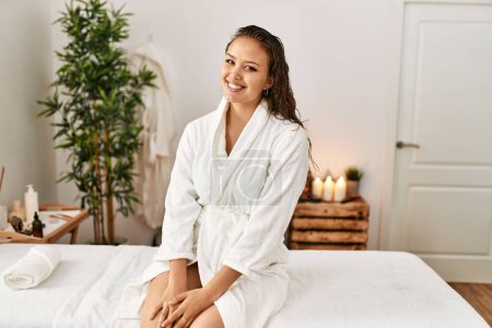 Photo for Young beautiful hispanic woman wearing bathrobe sitting on massage board at beauty salon - Royalty Free Image