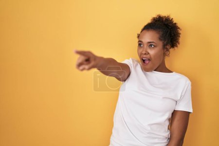 Photo for Young hispanic woman with curly hair standing over yellow background pointing with finger surprised ahead, open mouth amazed expression, something on the front - Royalty Free Image