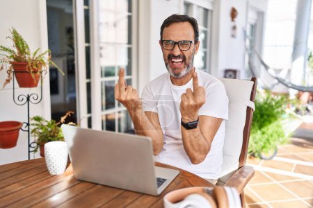 Foto de Middle age man using computer laptop at home showing middle finger doing fuck you bad expression, provocation and rude attitude. screaming excited - Imagen libre de derechos