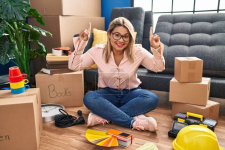 Foto de Young hispanic woman moving to a new home sitting on the floor smiling amazed and surprised and pointing up with fingers and raised arms. - Imagen libre de derechos
