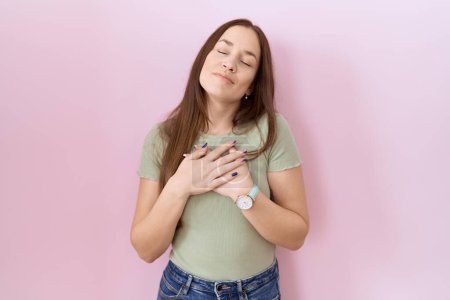 Téléchargez les photos : Beautiful brunette woman standing over pink background smiling with hands on chest with closed eyes and grateful gesture on face. health concept. - en image libre de droit