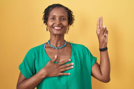 Téléchargez les photos : Femme africaine avec dreadlocks debout sur fond jaune souriant jurant avec la main sur la poitrine et les doigts vers le haut, faisant un serment de fidélité promesse - en image libre de droit