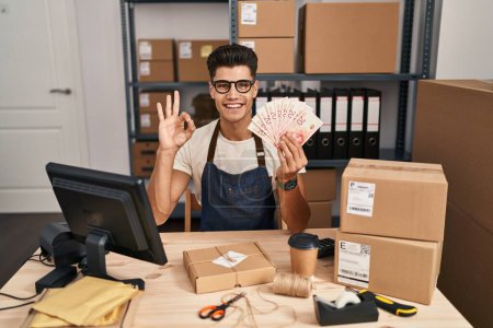Foto de Young hispanic man working at small business ecommerce holding shekels doing ok sign with fingers, smiling friendly gesturing excellent symbol - Imagen libre de derechos