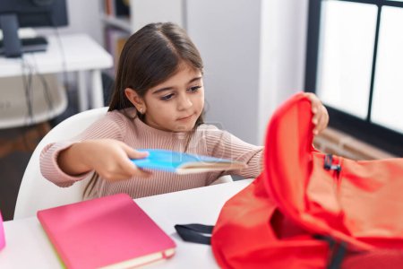Photo for Adorable hispanic girl student sitting on table putting book on backpack at classroom - Royalty Free Image