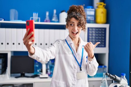 Téléchargez les photos : Hispanic woman with curly hair working at scientist laboratory doing selfie pointing thumb up to the side smiling happy with open mouth - en image libre de droit