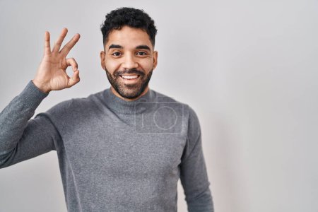 Foto de Hombre hispano con barba de pie sobre fondo blanco sonriendo positiva haciendo signo de ok con la mano y los dedos. expresión exitosa. - Imagen libre de derechos