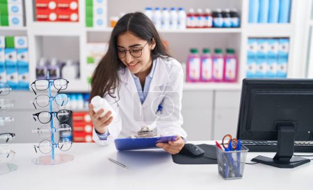 Foto de Joven chica hispana farmacéutica leyendo documento sosteniendo pastillas en la farmacia - Imagen libre de derechos