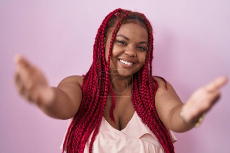 Téléchargez les photos : Femme afro-américaine avec les cheveux tressés debout sur fond rose regardant la caméra souriant à bras ouverts pour étreindre. expression joyeuse embrassant le bonheur. - en image libre de droit
