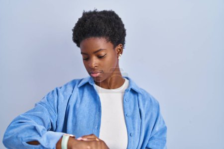 Téléchargez les photos : Femme afro-américaine debout sur fond bleu vérifier l'heure sur montre-bracelet, détendu et confiant - en image libre de droit