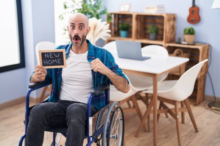 Téléchargez les photos : Hispanic man with beard sitting on wheelchair at new home scared and amazed with open mouth for surprise, disbelief face - en image libre de droit