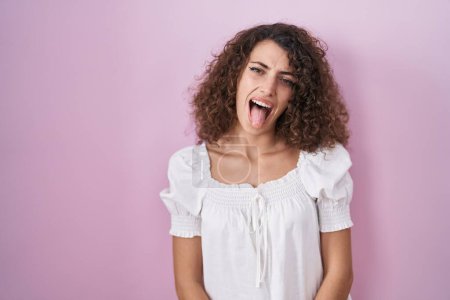 Photo for Hispanic woman with curly hair standing over pink background sticking tongue out happy with funny expression. emotion concept. - Royalty Free Image