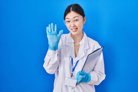 Téléchargez les photos : Chinese young woman working at scientist laboratory waiving saying hello happy and smiling, friendly welcome gesture - en image libre de droit